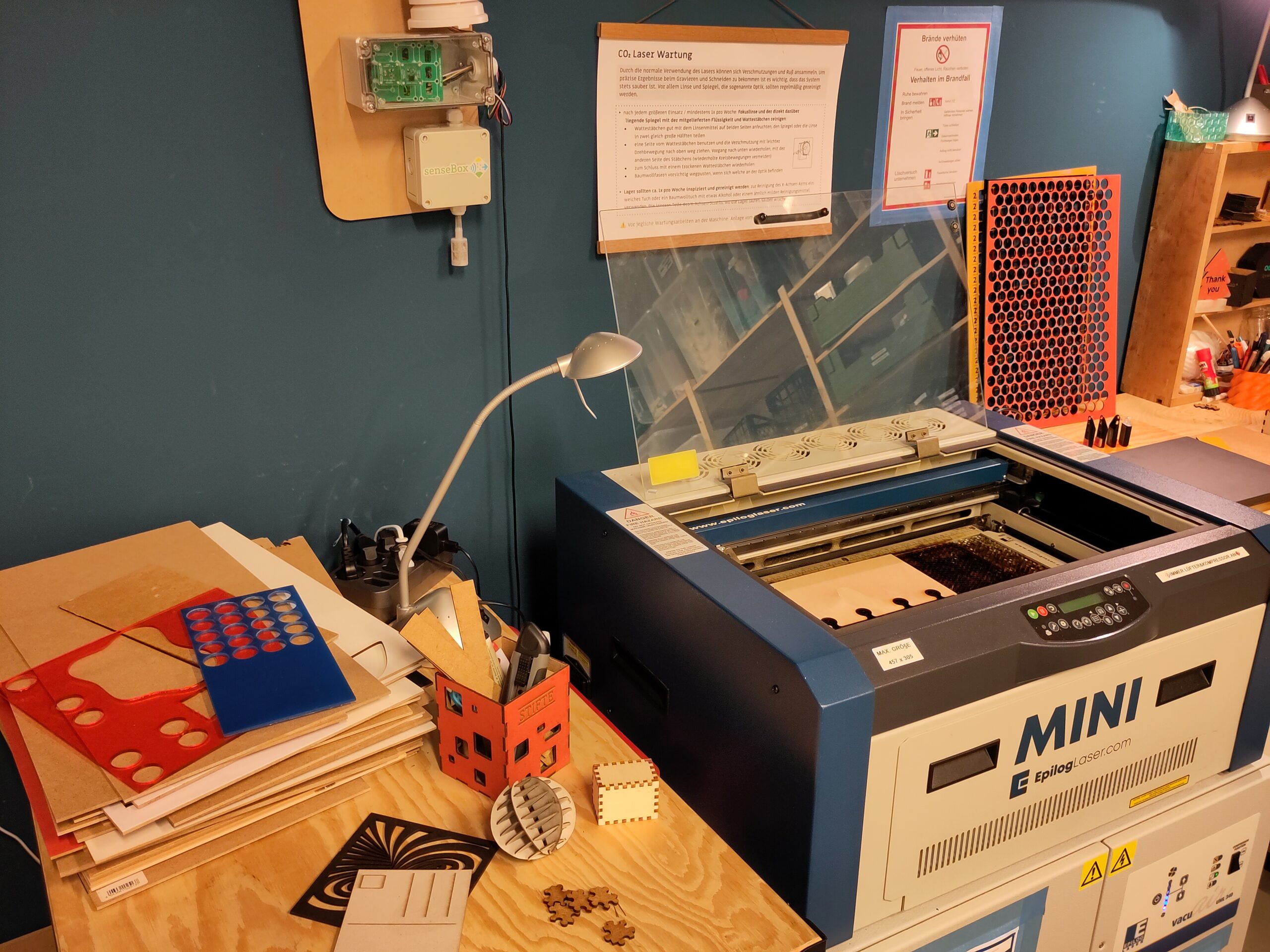 Ein Lasercutter in einer Werkstatt. Daneben ein Tisch mit verschiedenen Materialien