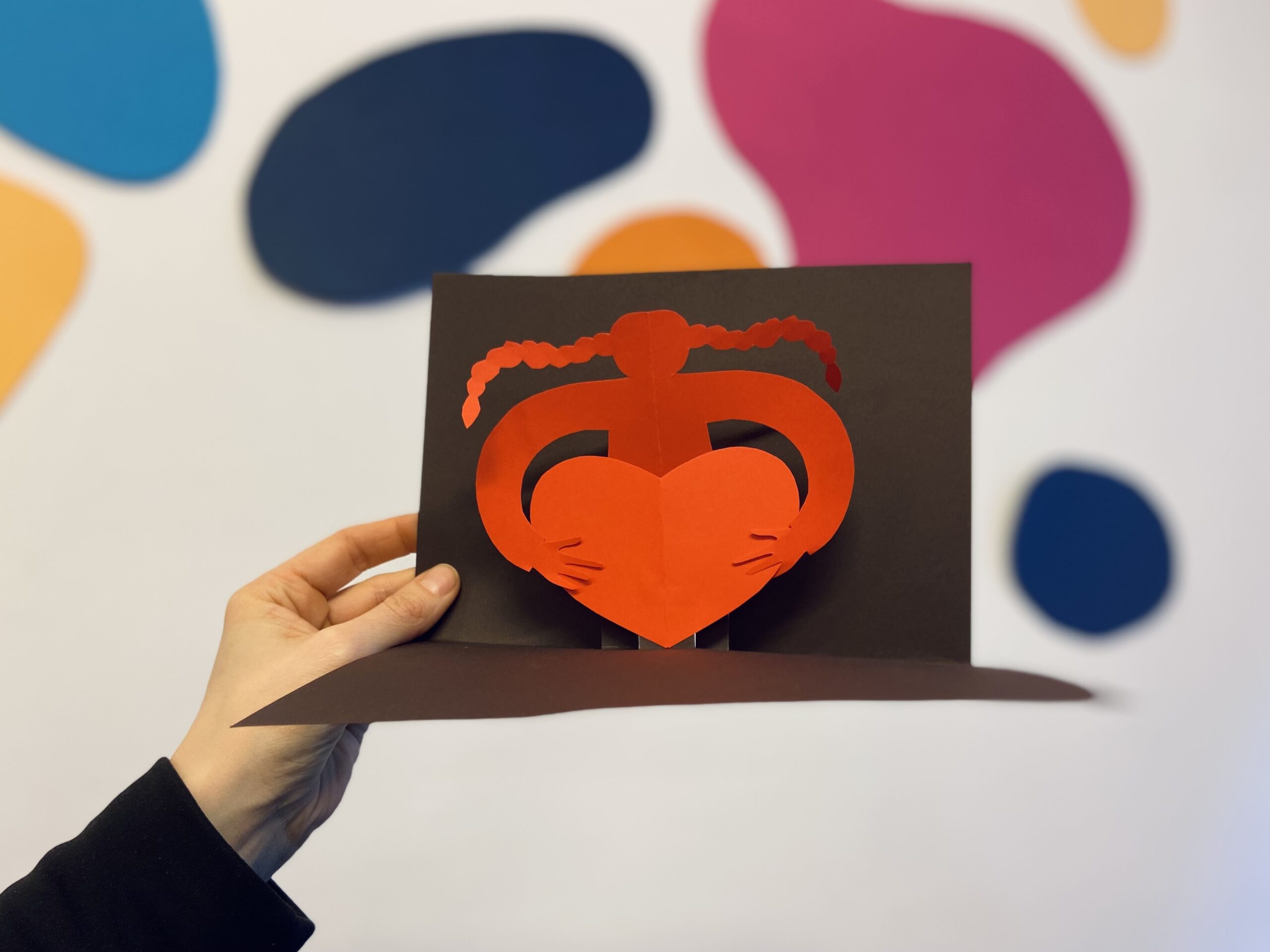 Eine Hand hält eine aufgefaltete Klappkarte. Darin ist ein aus rotem Papier geschnittene rote Figur mit langen Zöpfen, die ein Herz umarmt.