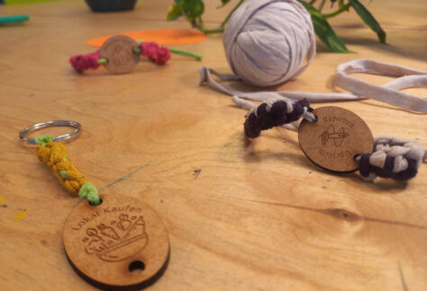 Auf einem Holztisch liegen selbstgeflochtene Armbänder und Anhänger mit kleinen Holzscheiben. Darauf sind Motive graviert und Sprüche wie 'Lokal kaufen' oder 'Reparieren'. Im Hintergrund liegt eine Rolle Garn aus einem alten T-Shirt